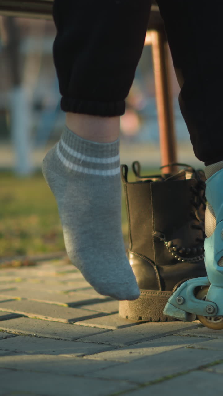 A person sitting in a park, with one leg in a rollerblade, pulling off a black boot from the other leg and preparing to wear the second rollerblade