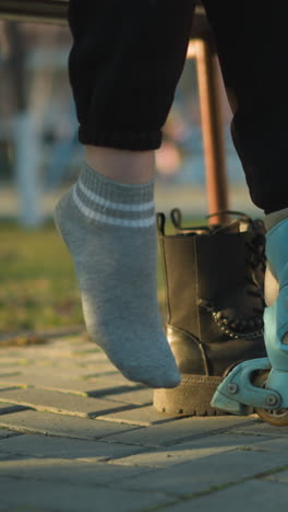 a person sitting in a park, with one leg in a rollerblade, pulling off a black boot from the other leg and preparing to wear the second rollerblade