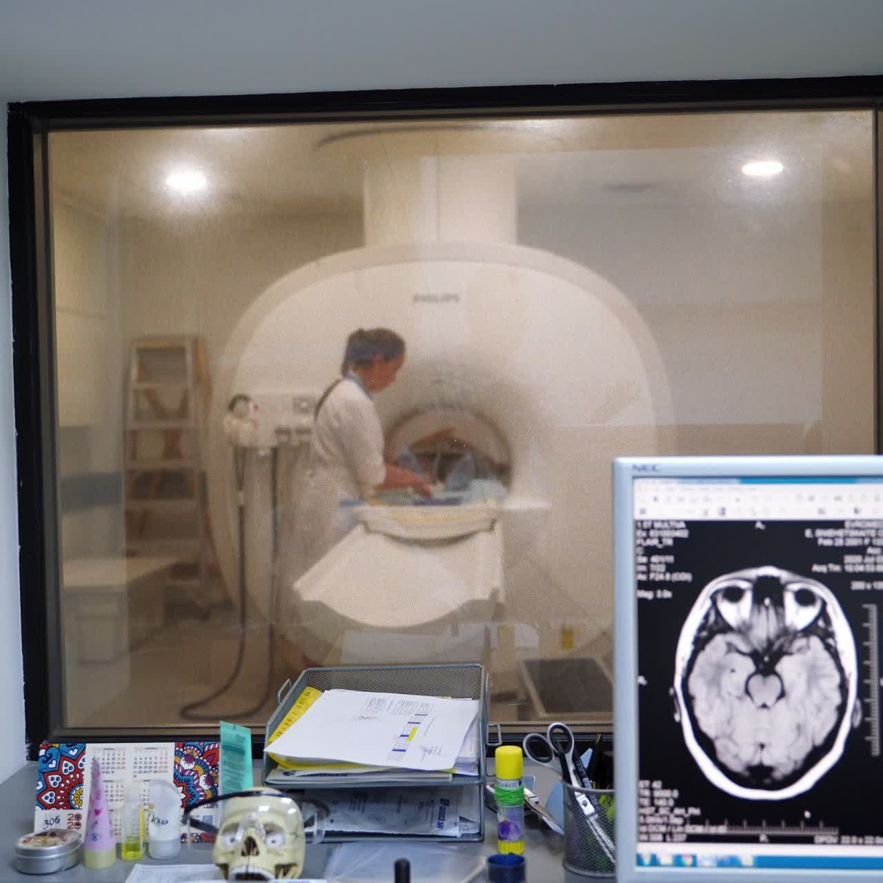 Looking through the window to the room with MRI scanning machine. Female nurse working near equipment. Computer screens with brain scans at foreground