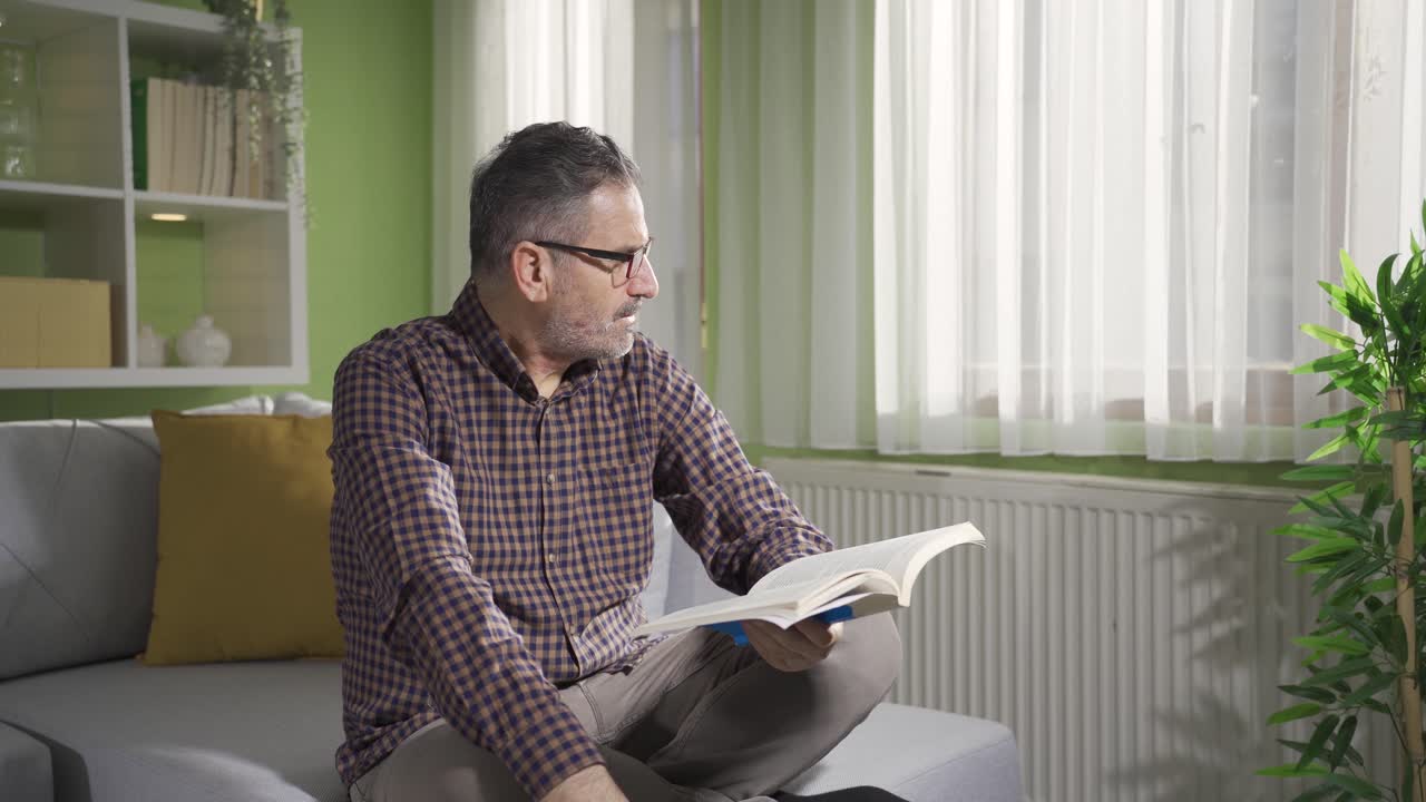 un hombre barbudo con gafas está leyendo un libro junto a la ventana.