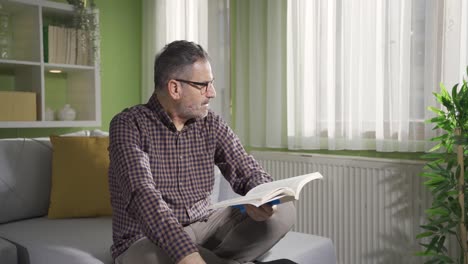 un hombre barbudo con gafas está leyendo un libro junto a la ventana.