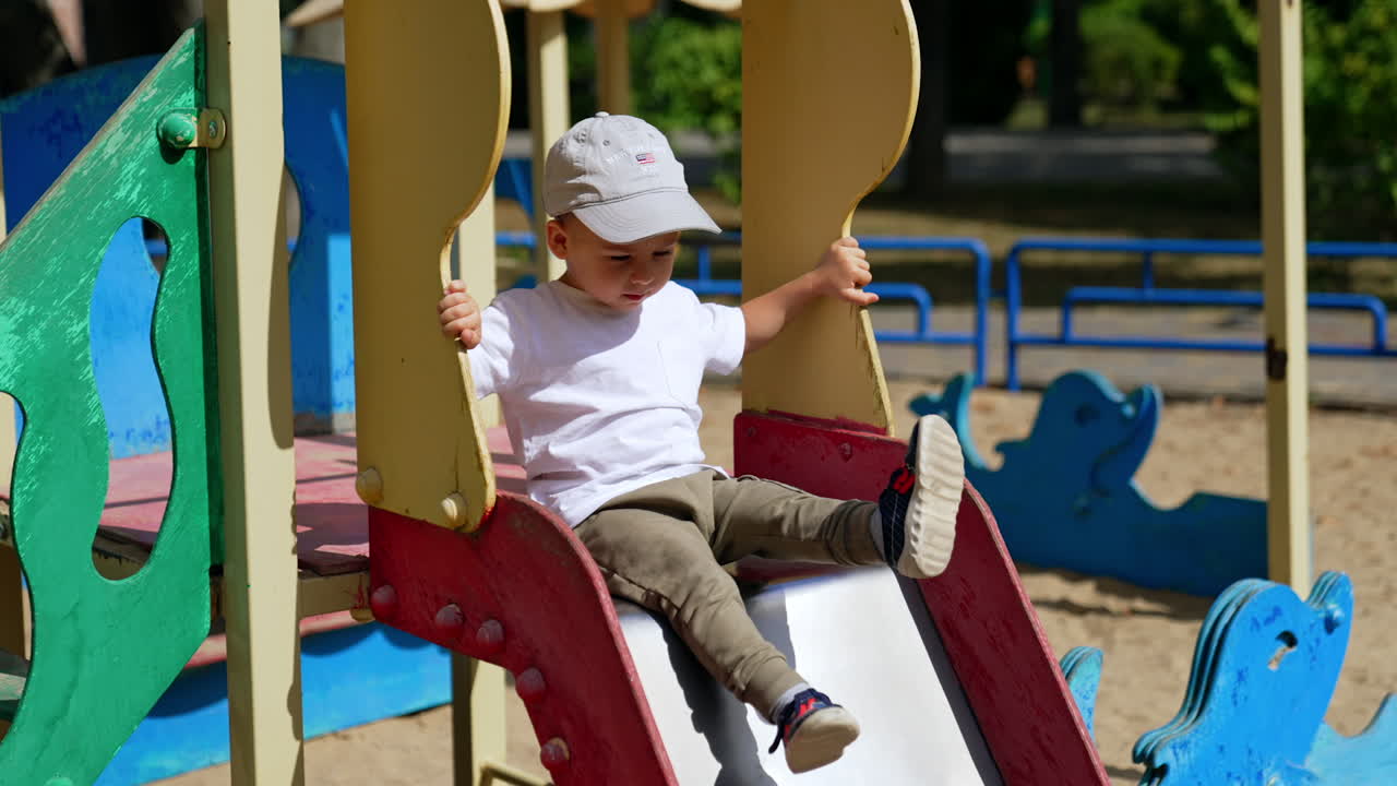 Energetic baby sitting on the slide on sunny day. Kid in cap bangs his feet by the slide on the playground.