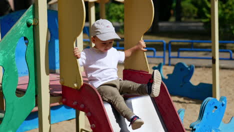 energetic baby sitting on the slide on sunny day. kid in cap bangs his feet by the slide on the playground.