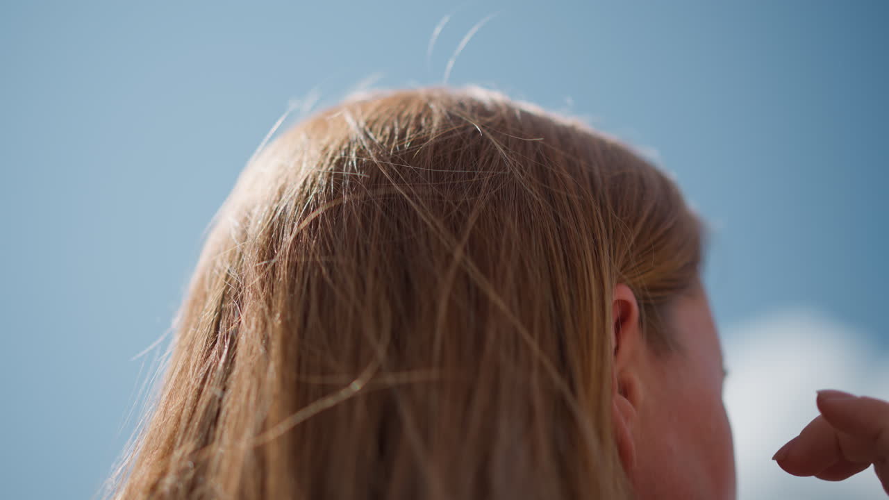 Rear close up of student with golden hair adjusting and scratching hair under bright sunlight, strands shining softly in breeze against clear blue sky, capturing warmth