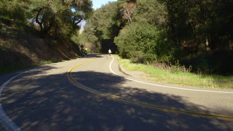 2 motocicletas conducen por un camino ventoso entre árboles verdes en california mientras un hombre camina solo en el fondo