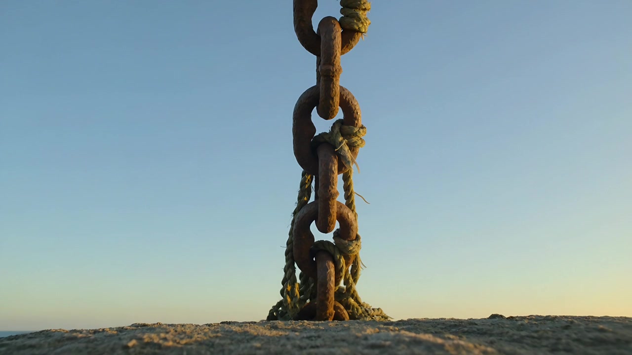 Swaying from gentle breeze, rusty metal chain rising along sea ledge with frayed rope and seaweed