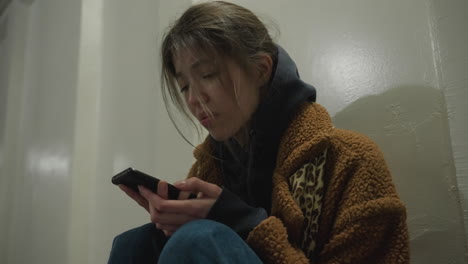 a close-up shot of a girl in a brown coat, sitting bent over in an underpass tunnel, holding her phone with a grieving expression, shaking her head in distress