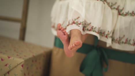 linda chica vestida con un vestido festivo sentada en regalos de navidad balanceando sus pies - primer plano