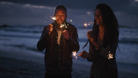 Una-Joven-Pareja-Multiétnica-En-La-Playa-Agitando-Bengalas-Celebrando-La-Víspera-De-Año-Nuevo-Disfrutando-De-Una-Velada-Divertida-Juntos-En-Un-Tranquilo-Fondo-Marino-Junto-Al-Mar-Por-La-Noche