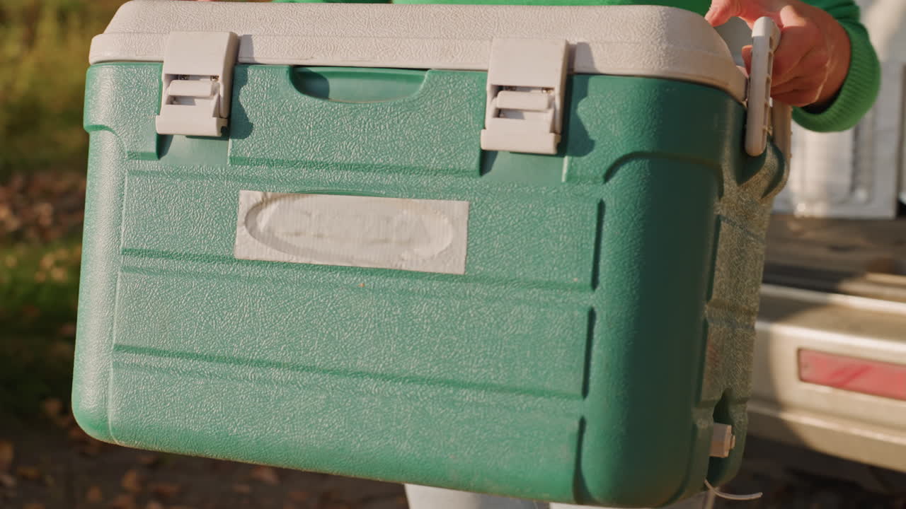 Frigo verde sollevato dal bagagliaio dell'auto per l'allestimento del campeggio, donna con maglione verde e jeans che trascina il frigo attraverso il campeggio coperto di foglie, primo piano su maniglie e plastica texturizzata, scatto d'azione di preparazione