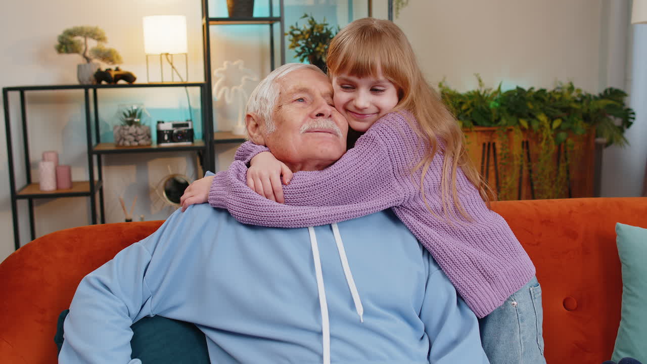 Allegra e affettuosa nipotina che abbraccia e bacia un nonno felice seduto sul divano di casa