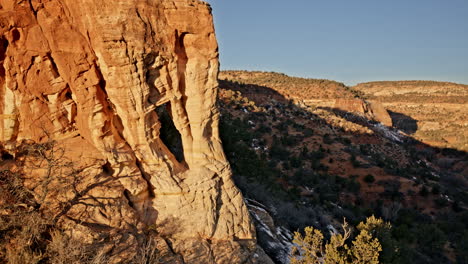 drone pans across a rugged red rock landscape, focusing on a striking natural arch at dawn.