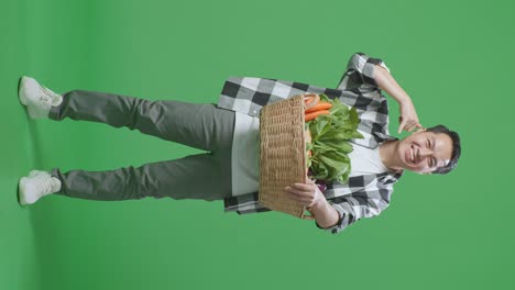 cuerpo lleno de granjero masculino asiático con canasta de verduras sonriendo cálidamente y haciendo hablar por teléfono gesto en el fondo de la pantalla verde en el estudio