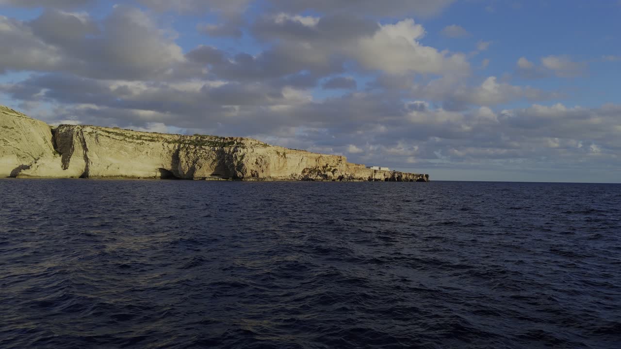 A fast drone flyover skims above the calm sea, revealing the Xaqqa Cliffs glowing in warm sunset light under a bright sky with soft clouds