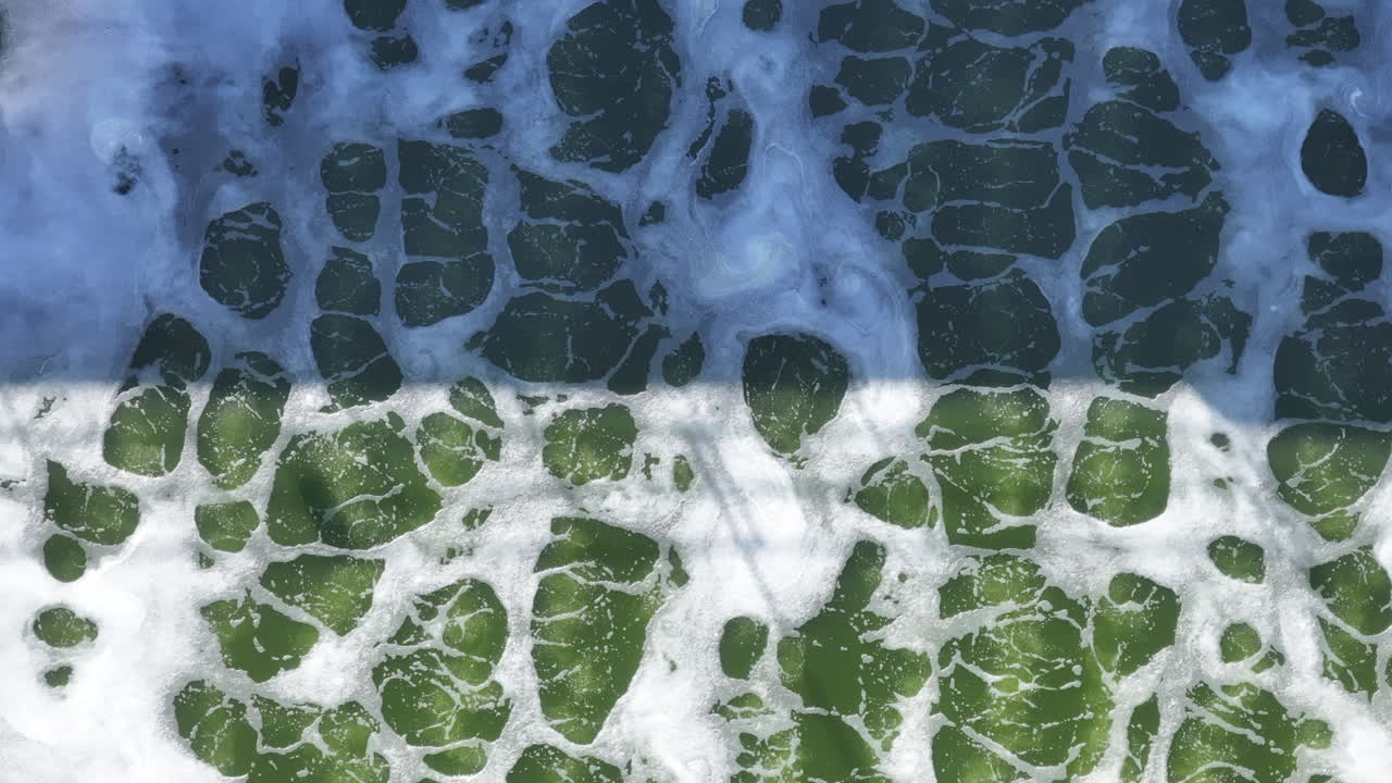 A zoomed aerial view of green, foamy water reveals rich bubble textures and movement under strong sunlight