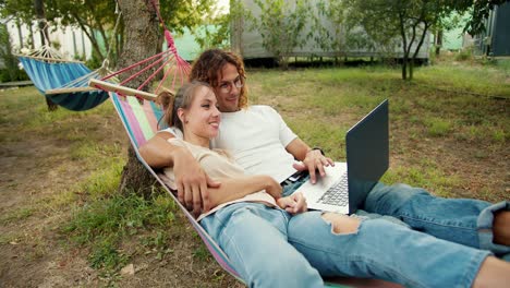 A-guy-in-glasses-with-curly-hair-and-his-girlfriend-are-lying-in-a-hammock-and-watching-a-video-on-a-laptop.-Rest-in-the-country-house