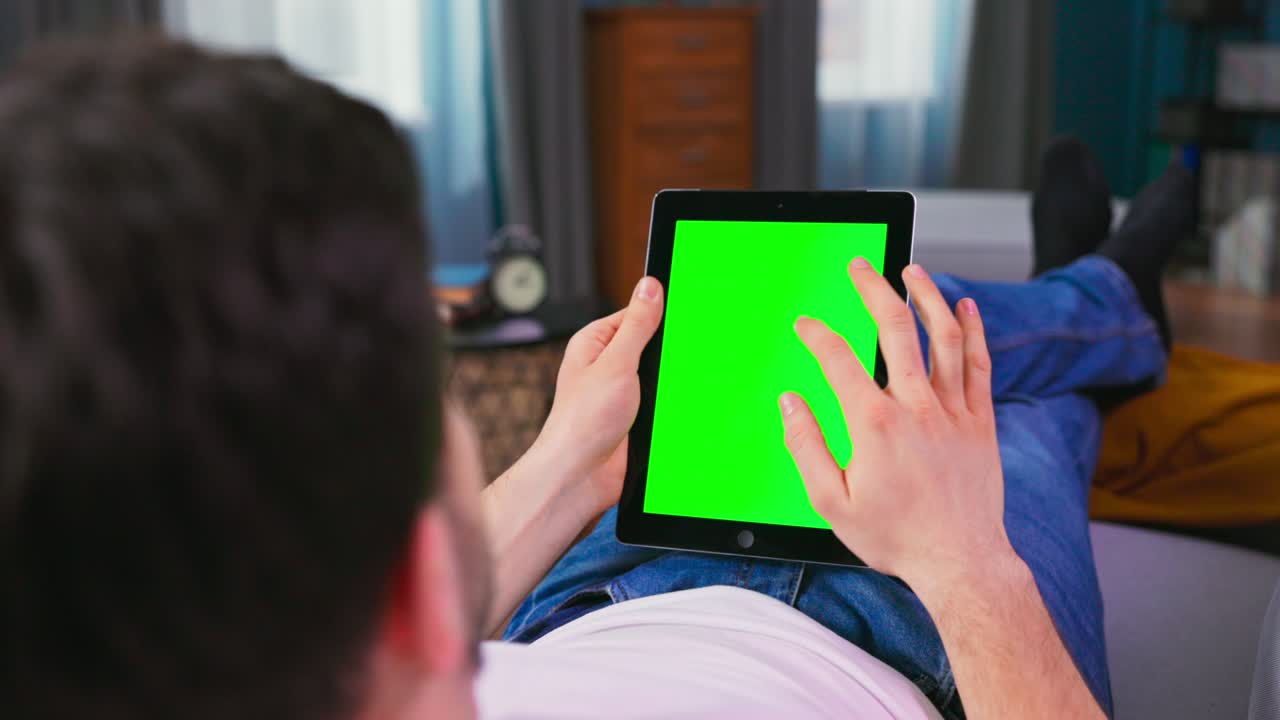 Young man at Home Resting on a Couch Using with Green Mock-up Screen Tablet Computer in Vertical Portrait Mode. Boy Using Gestures with Touchscreen Device, Browsing Internet, Watching Content
