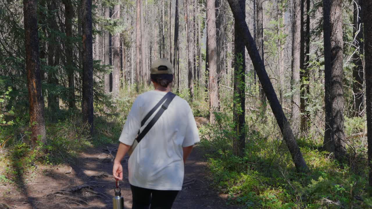 Woman walking a path through a pine forest, carrying a water bottle