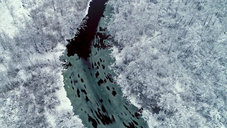 Winterluftaufnahme-Eines-Waldes,-Der-Mit-Einer-Dichten-Schicht-Aus-Weißem-Schnee-Und-Einem-Teilweise-Gefrorenen,-Gefrorenen-Fluss-Bedeckt-Ist