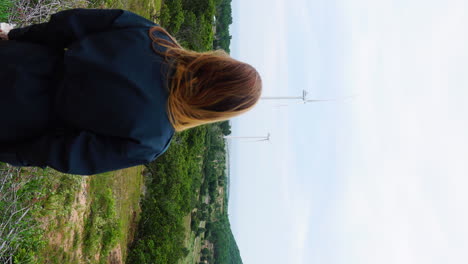 una chica de cabello pelirrojo parada en la cima de una colina y disfrutando de la vista de la montaña con una granja de molinos de viento en el fondo