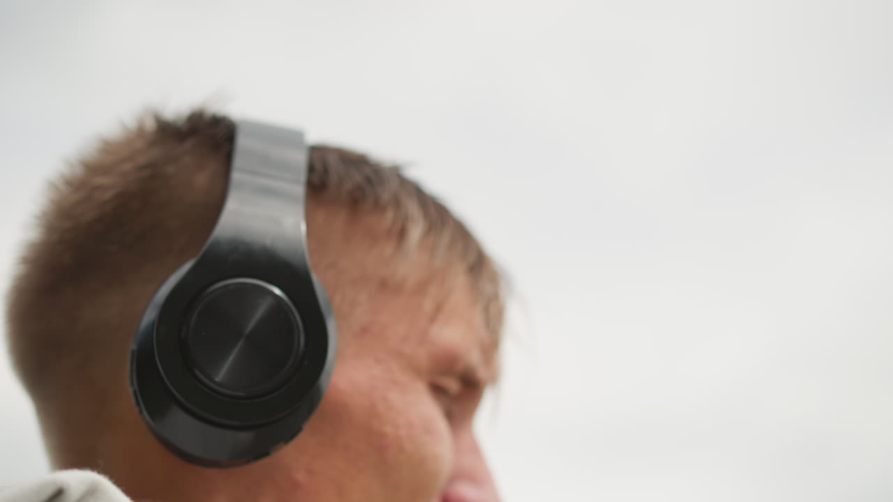 entrenamiento en una azotea nublada, hombre caucásico escuchando, sudor en la piel, gotas de lluvia en la sudadera con capucha, mandíbula apretada, mirada concentrada, ajuste táctil de los auriculares, cielo nublado, telón de fondo urbano y arenoso, entrenamiento lofi