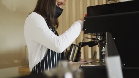 Caucasian-waitress-wearing-face-mask-standing-at-coffee-machine,-preparing-froth-milk