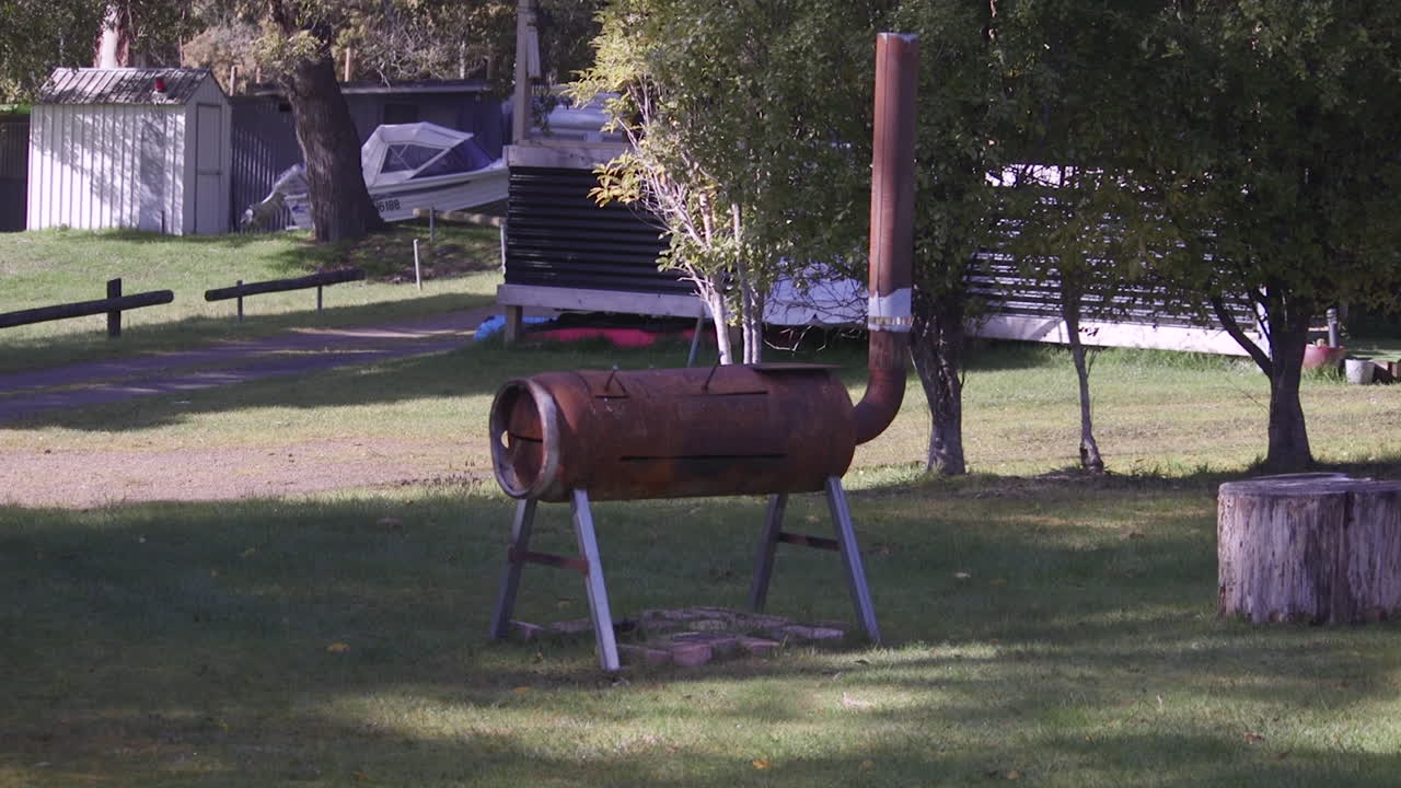 A metal fire pit grill used for outdoor cooking stands in a grassy area. Nearby trees provide shade, while a few boats are visible in the background. This spot is perfect for barbecues and gatherings.