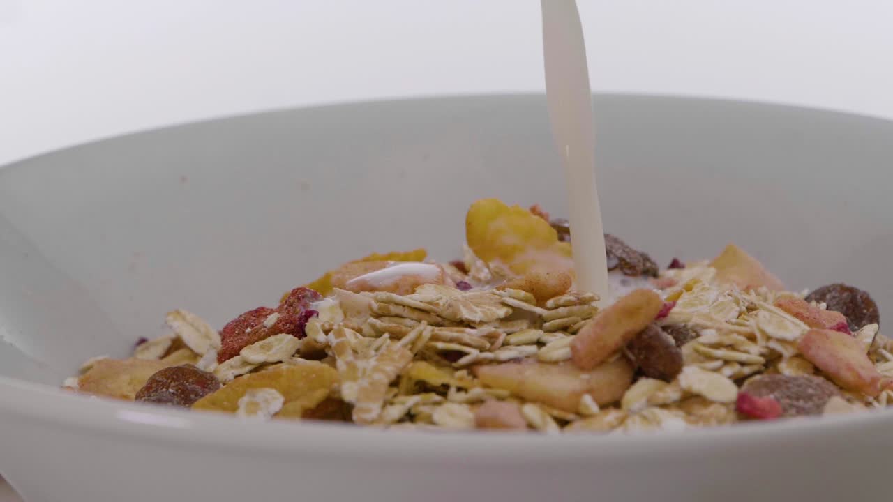 primer plano de un cuenco giratorio blanco sobre fondo blanco lleno de cereal vegano con frutas que se vierten en la leche de avena vegana para el desayuno