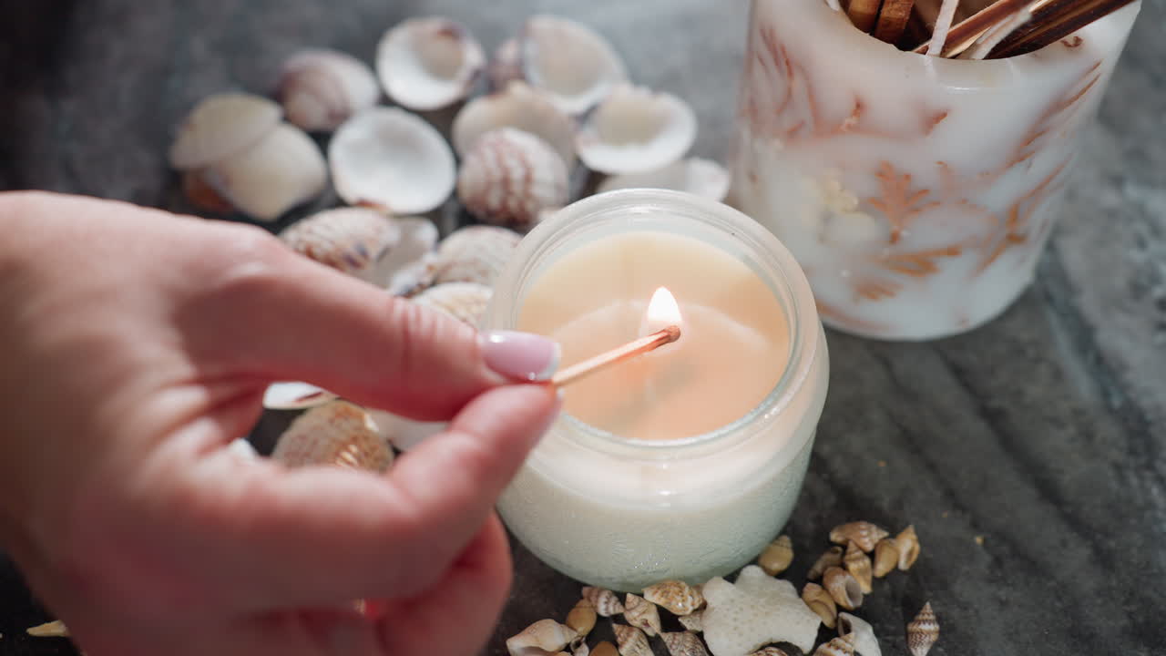 Close up of woman with polished nails holding burning matchstick near white candle surrounded by seashells, starfish, snail shells on marble surface