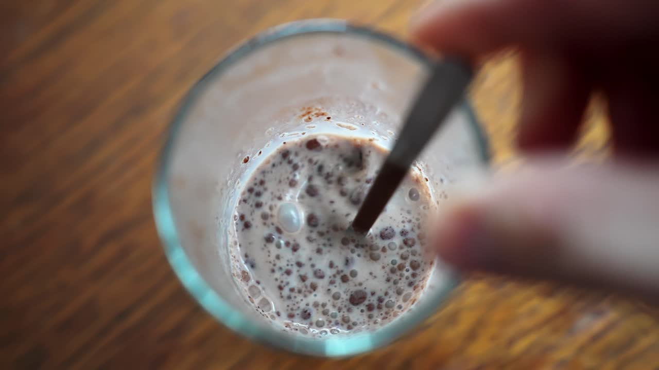 Primo piano dall'alto di una mano che mescola latte e cacao in un bicchiere, creando vortici spumosi su un tavolo di legno: preparazione casalinga e accogliente di cioccolata calda