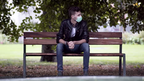 a young cool man in jeans, white t-shirt and a black leatherette jacket, wearing a green covid-19 facemask, sitting on a park bench under a tree beside a fountain, impatiently waiting for someone, 4k