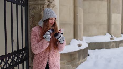 young woman in pink jacket texting on phone in snowy city