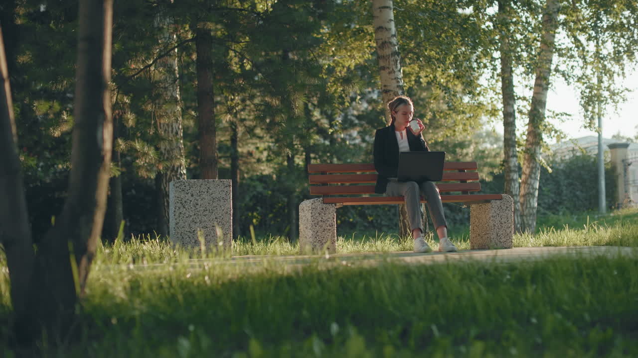 Freelancer seated outdoors on wooden bench working on system and sipping juice surrounded by lush greenery in park setting with warm morning light