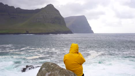 man with yellow waterproof puffer jacket looking at ocean and mountains in vidareidi, faroe islands