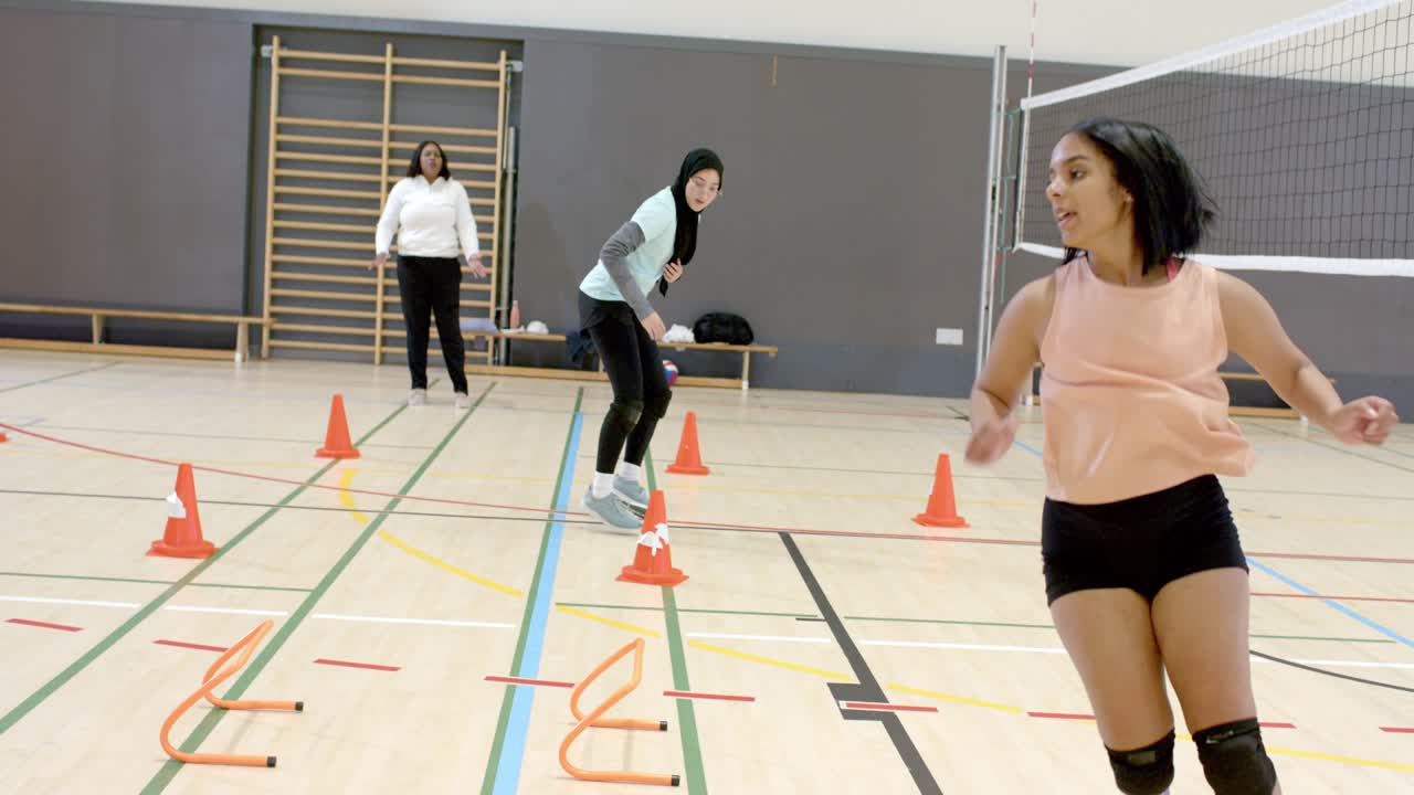 Coach initiating drill guiding diverse women's volleyball team performing hurdle-cone drills in gym