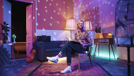 Young-stylish-caucasian-man-with-long-hair-and-glasses-sitting-on-a-chair-and-reading-a-book-in-a-retro-room-at-home