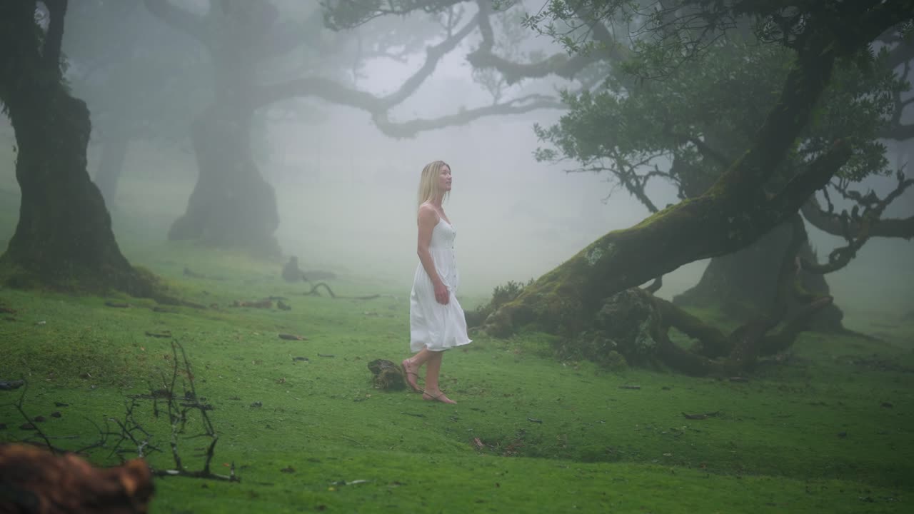 Attractive blond woman in angelic dress walks in eerie enchanted foggy forest