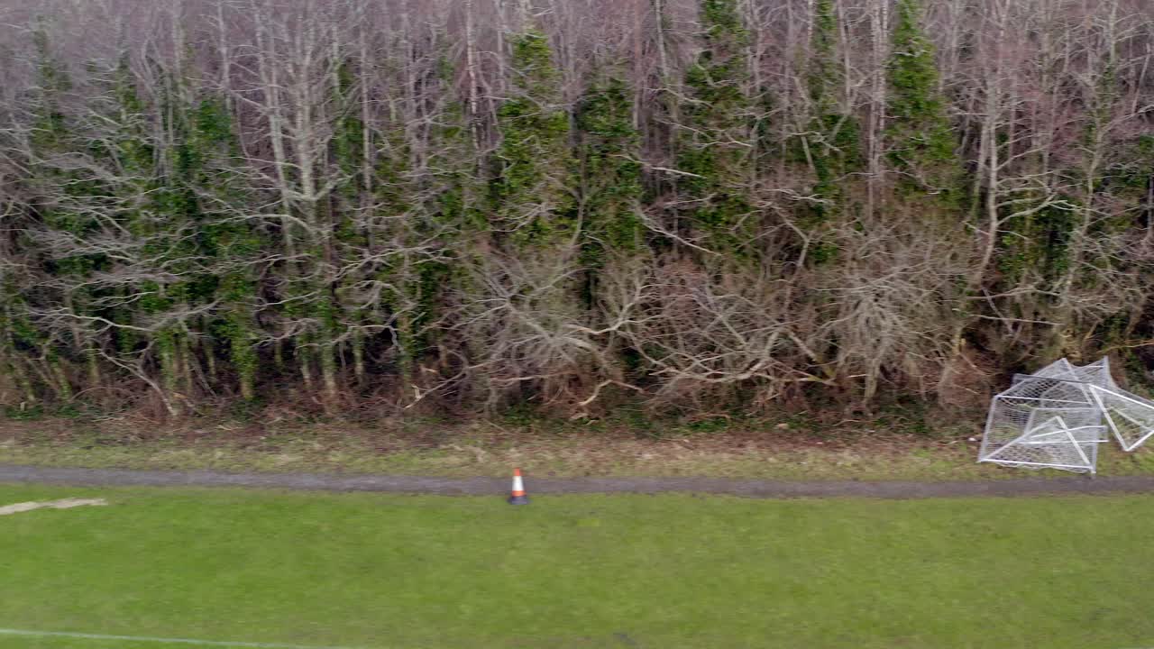 Aerial overview tracking left of damaged woodland with uprooted trees and scattered branches along path with broken football goals, Barna Woods, Galway Ireland