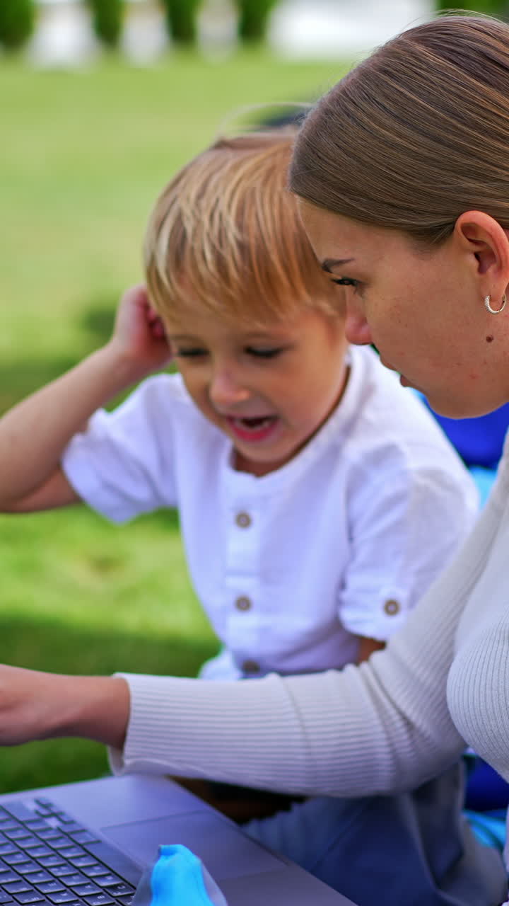Long-haired blonde woman and her little son sitting outdoors. Both look at computer discussing something cheerfully. Side view. Vertical video