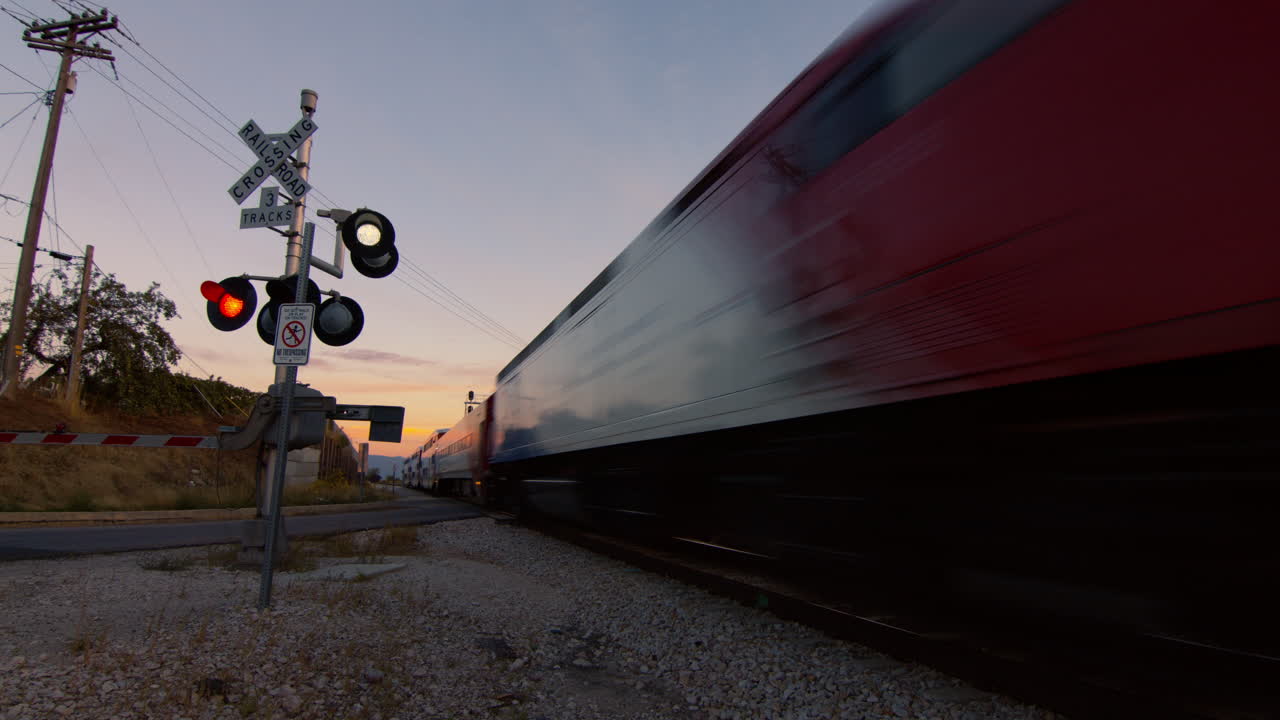 A railroad crossing at sunset.  The lights on the sign are flashing and a commuter train passes the camera.