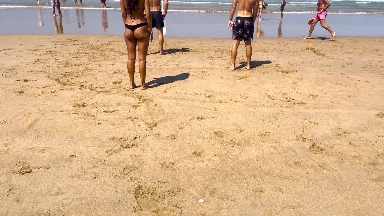 teenager woman in a black bikini playfully shakes sand off her hands onto her buttocks