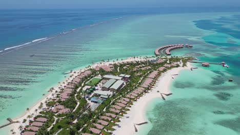 Luxury waterfront Bungalows at resort during sunny day in Maldives. Coral reef with turquoise ocean water. Aerial rising top down shot. Establishing shot.