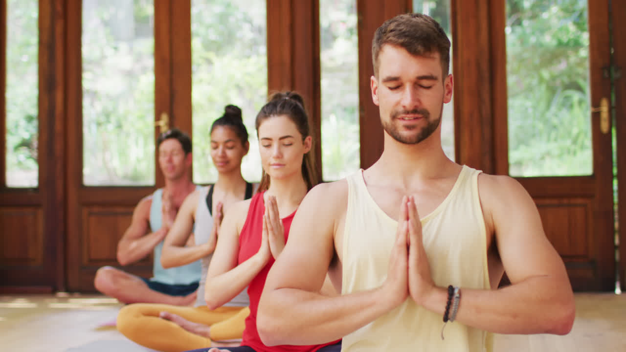 grupo diverso sonriente sentado en posición de yoga con los ojos cerrados, durante la clase de yoga en el estudio