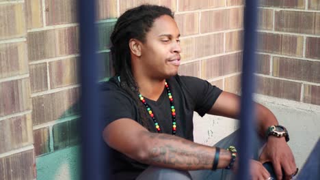 African American man with dreadlocks sitting on ground talking to someone and reacting, saying “you’re crazy”, and laughing in a friendly way