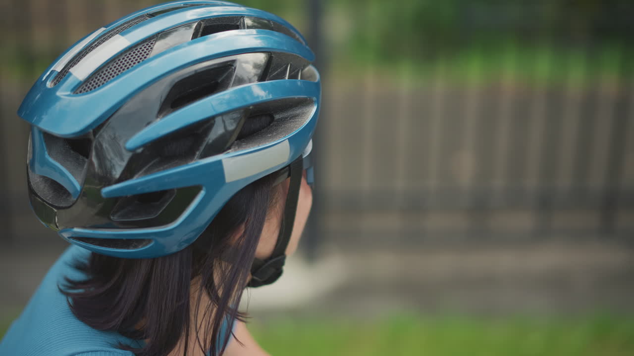 woman cyclist close up blue helmet, detailed vents and straps. reflective visor, urban fence background, light rain droplets, bike preparation and safety check, focused expression, textured shell