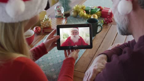 Kaukasisches-Paar-Mit-Weihnachtsmützen-Nutzt-Tablet-Für-Weihnachtsvideoanruf-Mit-Weihnachtsmann-Auf-Dem-Bildschirm