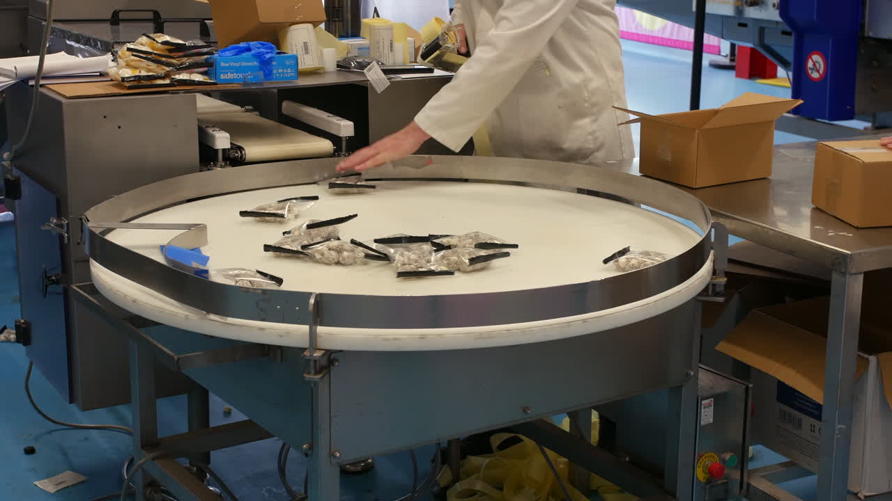 A conveyor table in a sweet factory displays packaged confectionery rotating toward a worker in a white coat. The worker holds a tape dispenser and packs products into open cardboard boxes