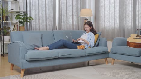 full body of asian teen girl eating snacks and typing on a laptop while lying on sofa in the living room at home