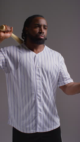 Vertical-Video-Studio-Portrait-Of-Serious-Male-Baseball-Player-Wearing-Team-Shirt-Holding-Bat-And-Throwing-Ball-In-The-Air-Shot-Against-Grey-Background