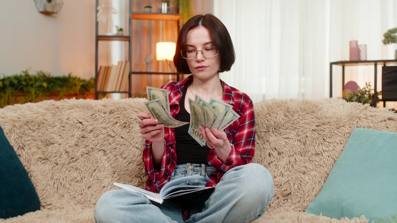 Seorang wanita muda menulis anggaran dalam buku catatan di rumah dengan ekspresi fokus sambil menghitung uang kertas dolar